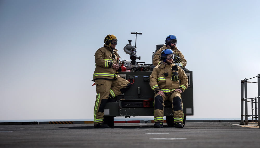 3 Navy sailors in fire protective clothing lean and sit of a piece of equipment on board a ship