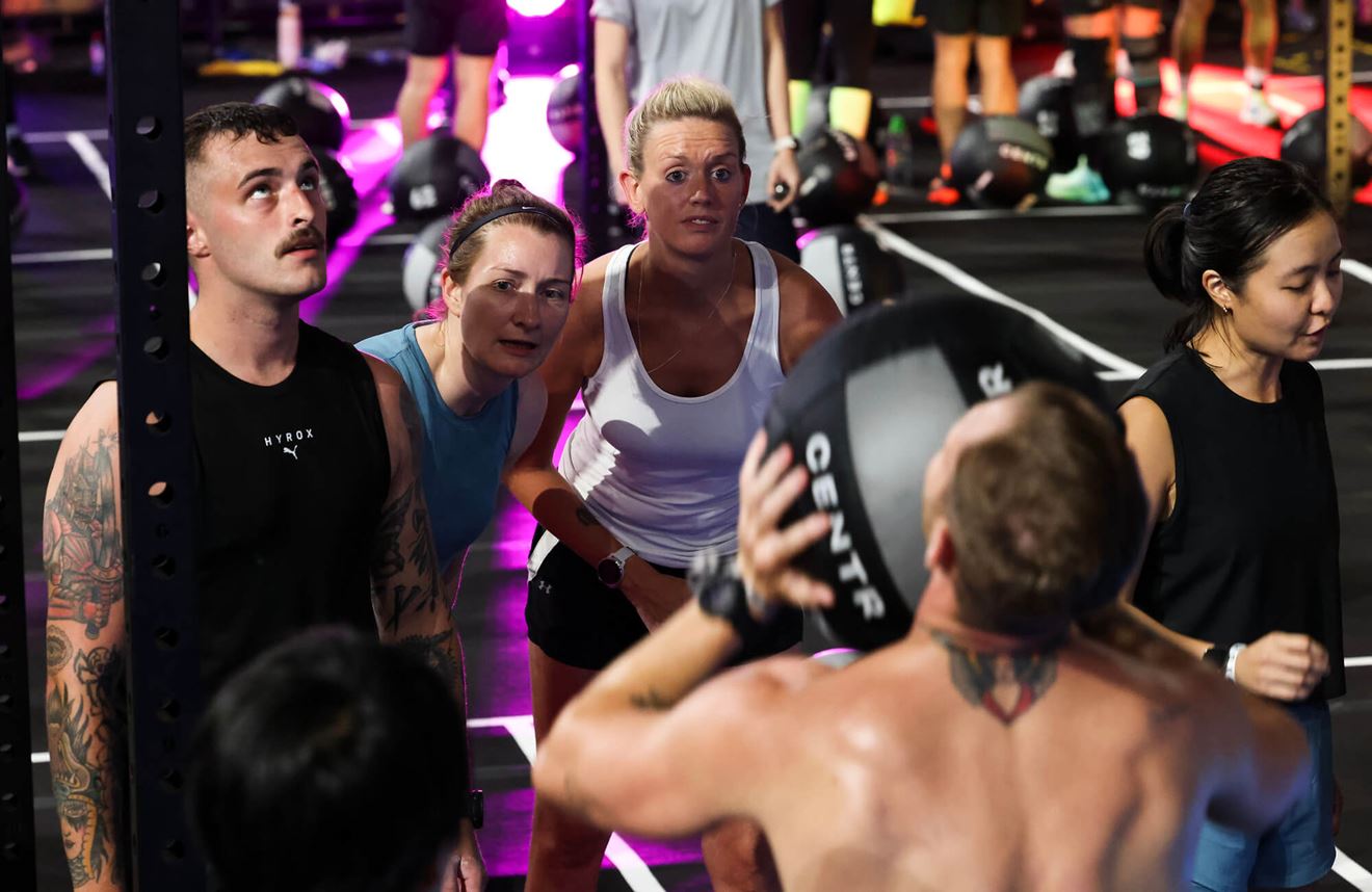 A colour photo shows a group of Royal Navy service members work out together in a fully-equipped Royal Navy gym. One service member lifts a large medical ball weight as his colleagues look on.