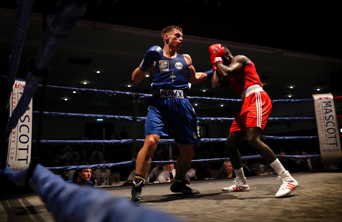 A colour photos shows a Royal Navy boxer mid-fight. He swings as his opponent blocks a hit to the face. In the shadows of the background, a few onlookers can be seen.