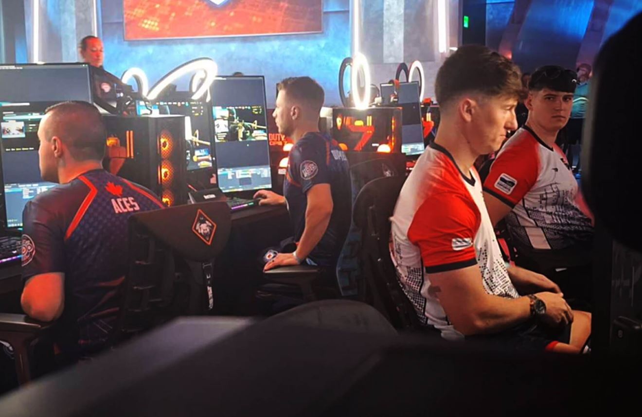 A colour photo shows two Royal Navy eSports players on-break during a tournament. Sitting behind them are two players from the Canadian Armed Forces. In the background are several large monitors, computers, and ring lights.
