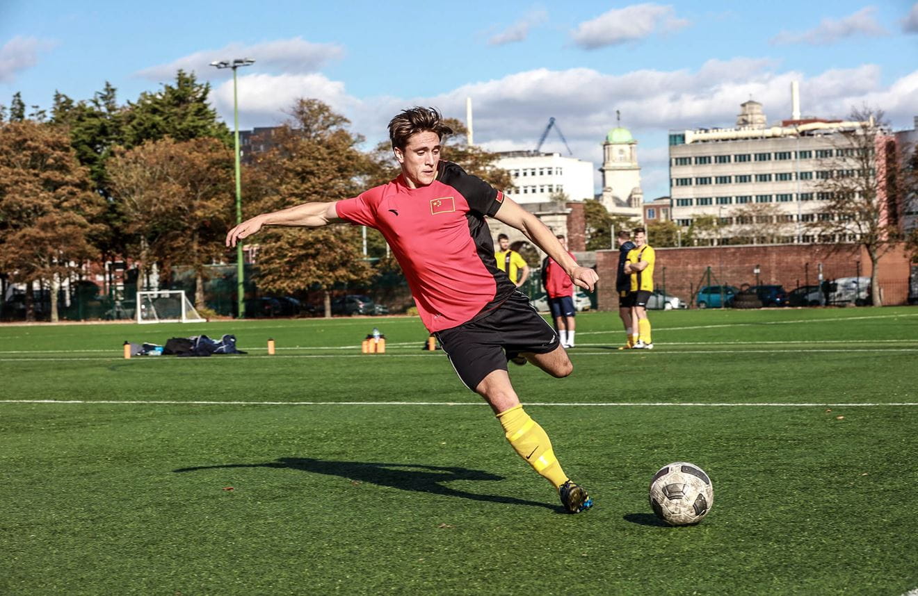 A colour photo shows a Royal Navy footballer on the pitch. He swings his leg back to kick a ball forward. In the background are several of his teammates and large buildings in the distance.