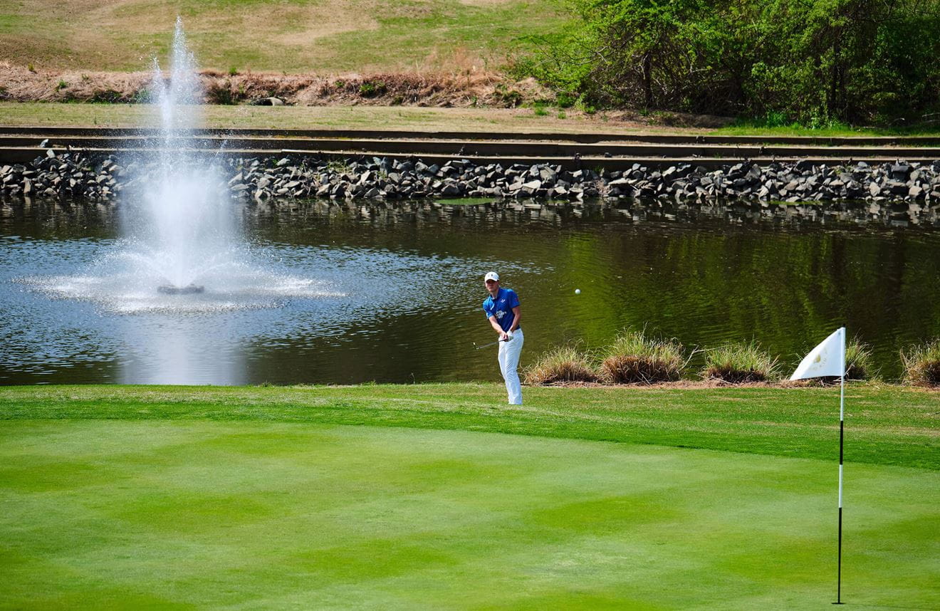 A colour image shows a wide angle of a Royal Navy golfer on course. He is swinging a club, as a ball heads towards a hole in the foreground. In the background is a large water fountain and pond.
