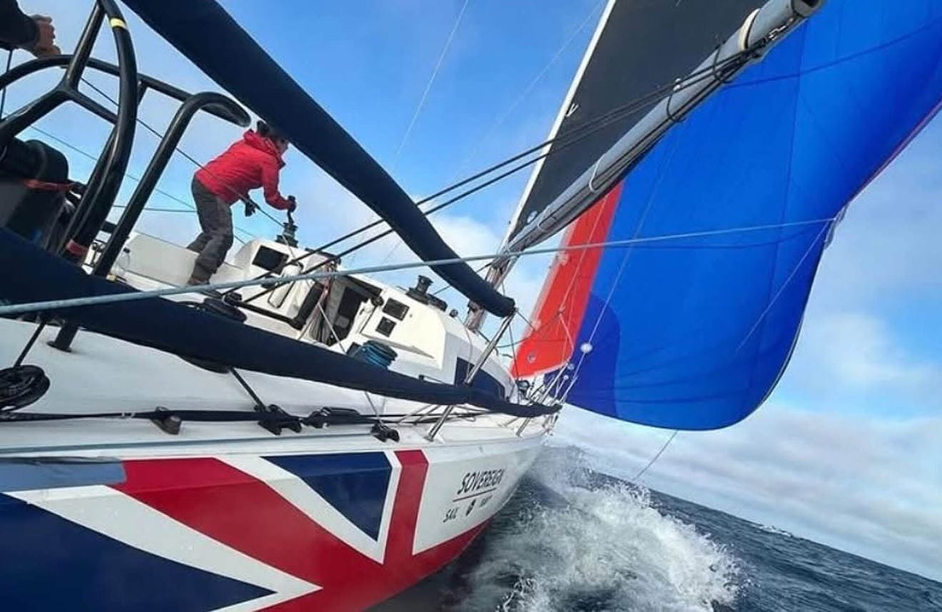 A dynamic colour photo shows a Royal Navy yacht racing through open waters. Its hull is painted in a Union Jack decal. One sailor mans the controls as their crew mate controls the sails. The water foams and splashes as the boat speeds through.
