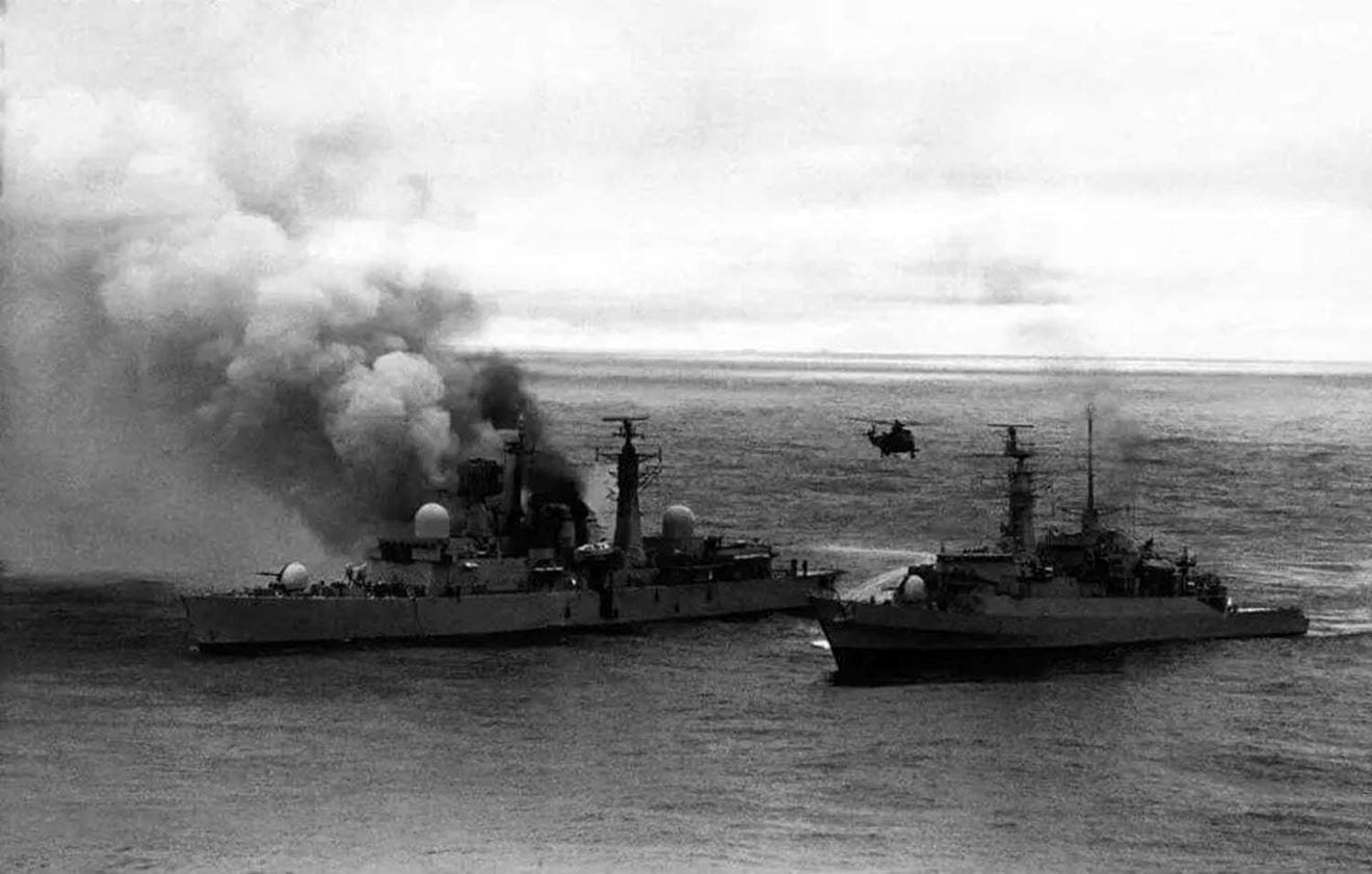HMS Arrow approaches the burning HMS Sheffield with a Sea King transferring casualties