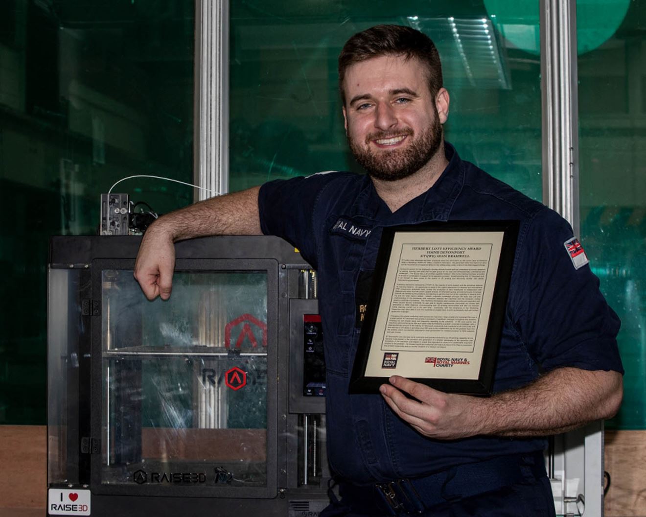 Engineering Technician Sean Bramwell with his recognition award for his efforts during the COVID response standing next to the 3D printer where he produced PPE components
