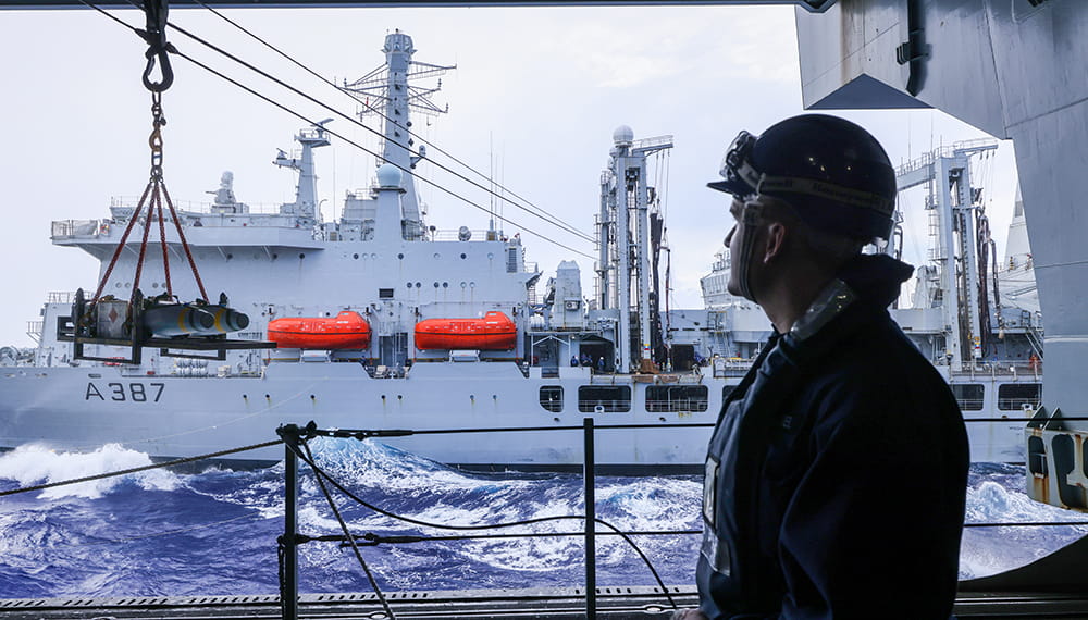 Royal Navy sailor in hardhat looks from the deck of one ship to another during a supply transfer