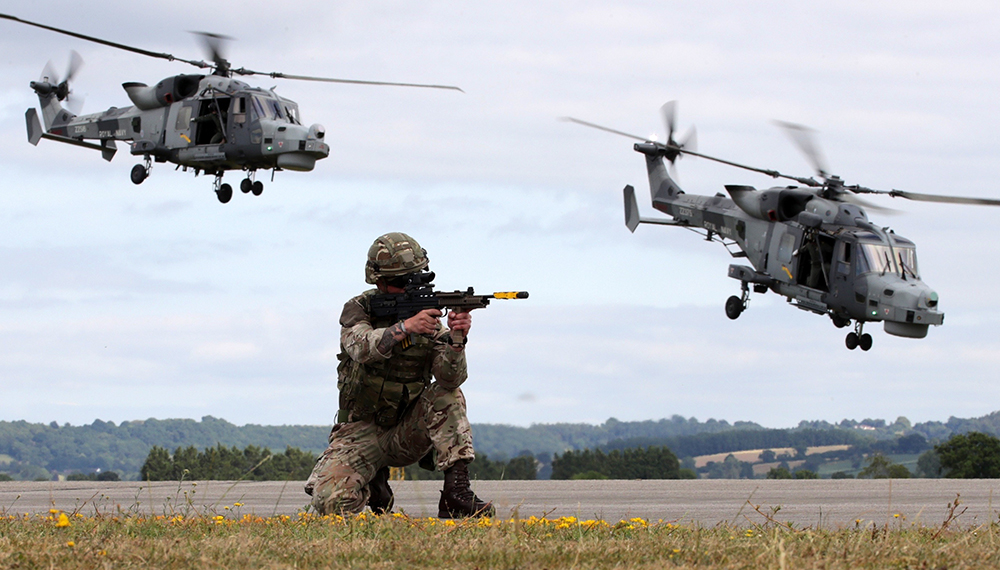 Royal-Navy-What-we-do-Respond-02-deploying-rapidly.jpg Royal Marine on one knee aims his gun while two two helicopters fly overhead