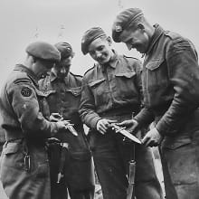 A black-and-white photo shows four Royal Marines of the 46 and 45 Commandos comparing battle trophies after the successful crossing of the Rhine at Wesel, Germany, 28 March 1945.