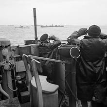 A black-and-white photo shows two British officers keeping watch at sea for German U-boats.