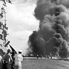 A black-and-white photo shows large, black clouds of smoke from the aftermath of a kamikaze strike on HMS Victorious. In the background, damage control parties use water hoses to control the fire.