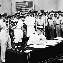 A black-and-white photo shows Royal Navy Admiral Sir Bruce Fraser signing the instrument of surrender with Japan, aboard USS Missouri in Tokyo Bay. In the background are several allied servicemen and an American flag hangs on the wall.