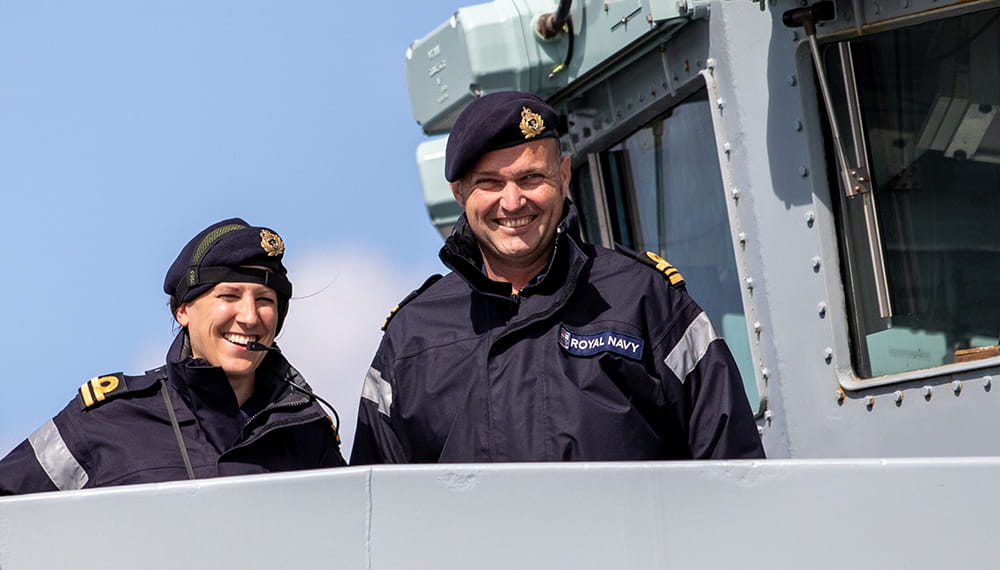 royal-navy-careers-joining-options-03 two smiling royal navy crewmembers in uniformed jackets on bridge of vessel smiling into camera in sunny conditions
