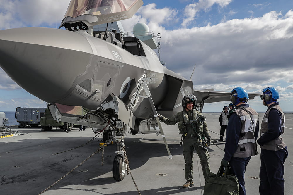 uniformed and blue helmeted fleet air arm aircrew preparing to board aircraft on board aircraft carrier under cloudy and blue sky
