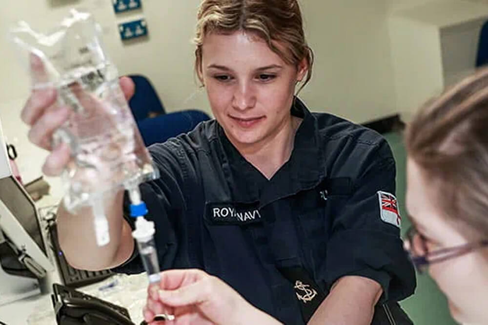 A colour photo shows a Royal Navy nurse preparing a medical I.V. bag. She is wearing a blue Royal Navy work uniform. A second medical assistant is cropped out of frame; she is assisting with the I.V.