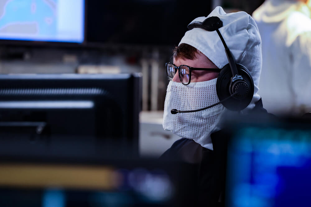 A colour image shows a Royal Navy warfare specialist sitting at a workstation. He is wearing glasses, protective anti-flash gear,  and a headset. In the blurred foreground are computer screens.