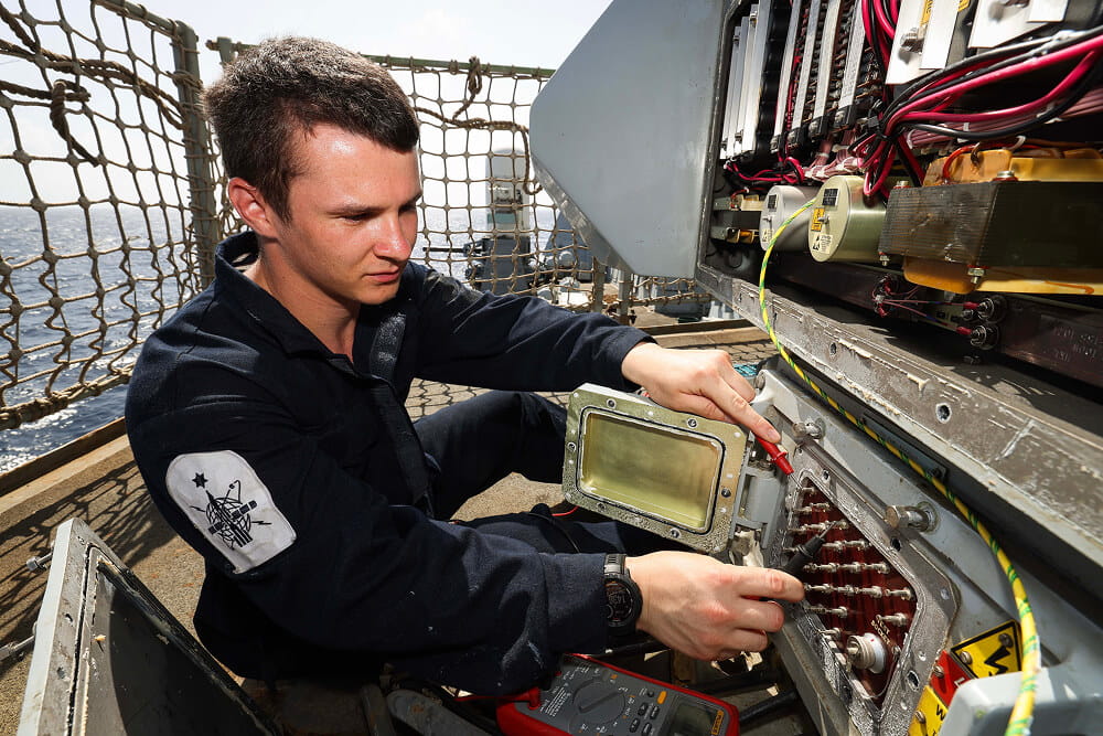 A colour image shows a Royal Navy engineer technician working on a large electrical board on the deck of a ship. There are several black and red cables, metal switches, and caution signs on the inside of the panel. He holds a pair of specialist tools in his hand and wears a blue Royal Navy work uniform. In the background are safety nets and a glimpse of the vast ocean behind him.