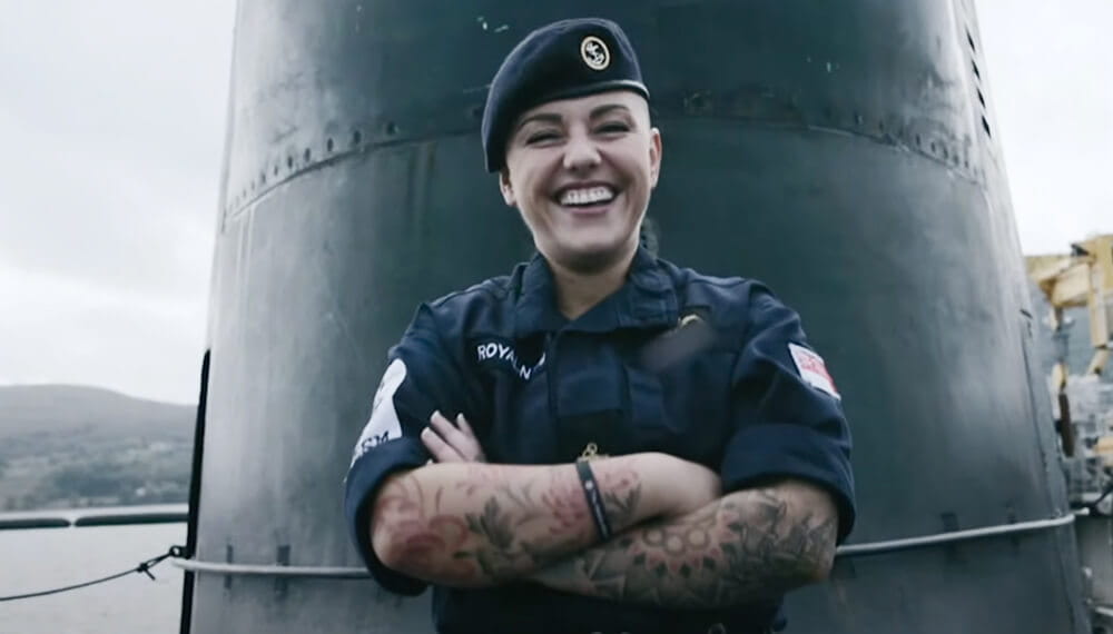 A submariner smiling with crossed arms in front of a submarine with uniform on