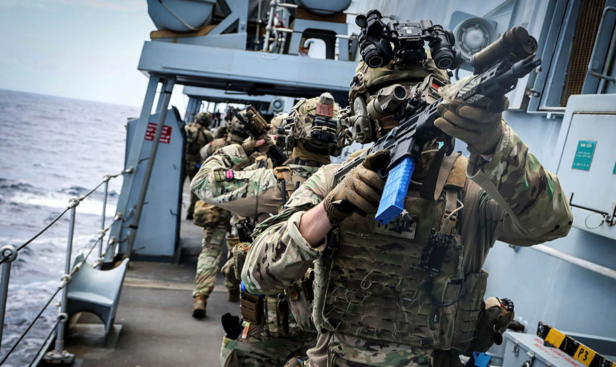 Several Royal Marines on the deck of a ship, wearing combat fatigues and carrying automatic firearms. A Commando aims his weapon to something out of frame