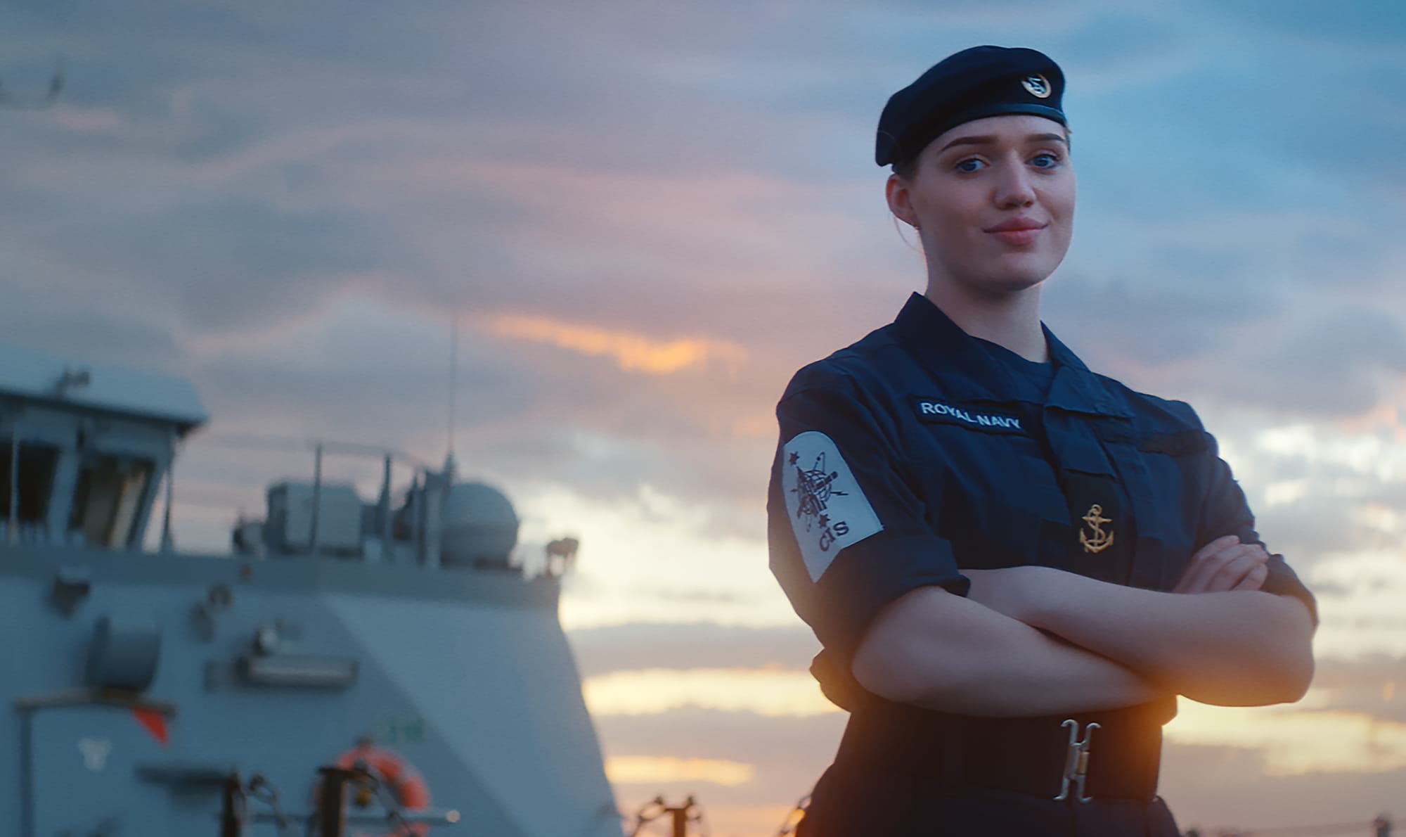A Royal Navy crew member wearing a blue beret stands on the deck proudly looking at the camera