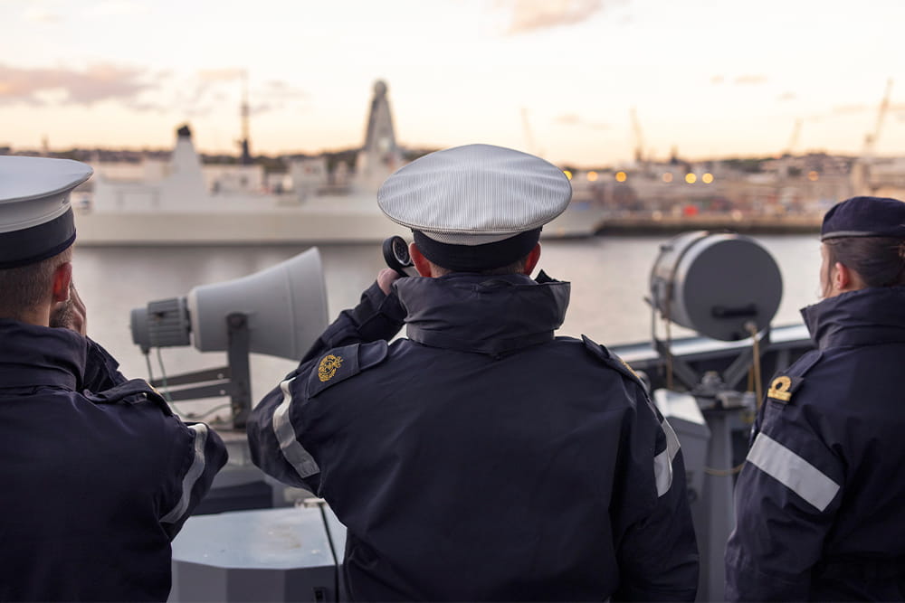 3 Navy personnel look off at another ship in the port, one looks at a compass