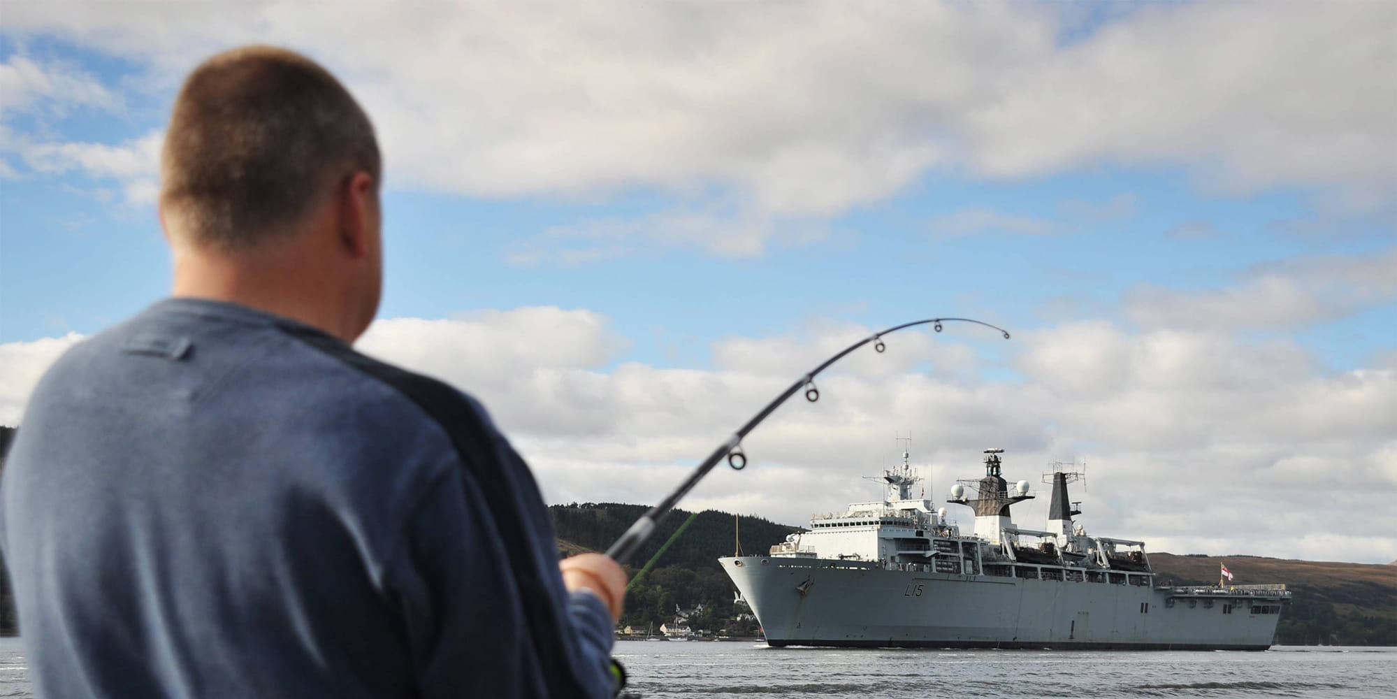 Fisherman casts a line into the sea with a Albion class ship in the background