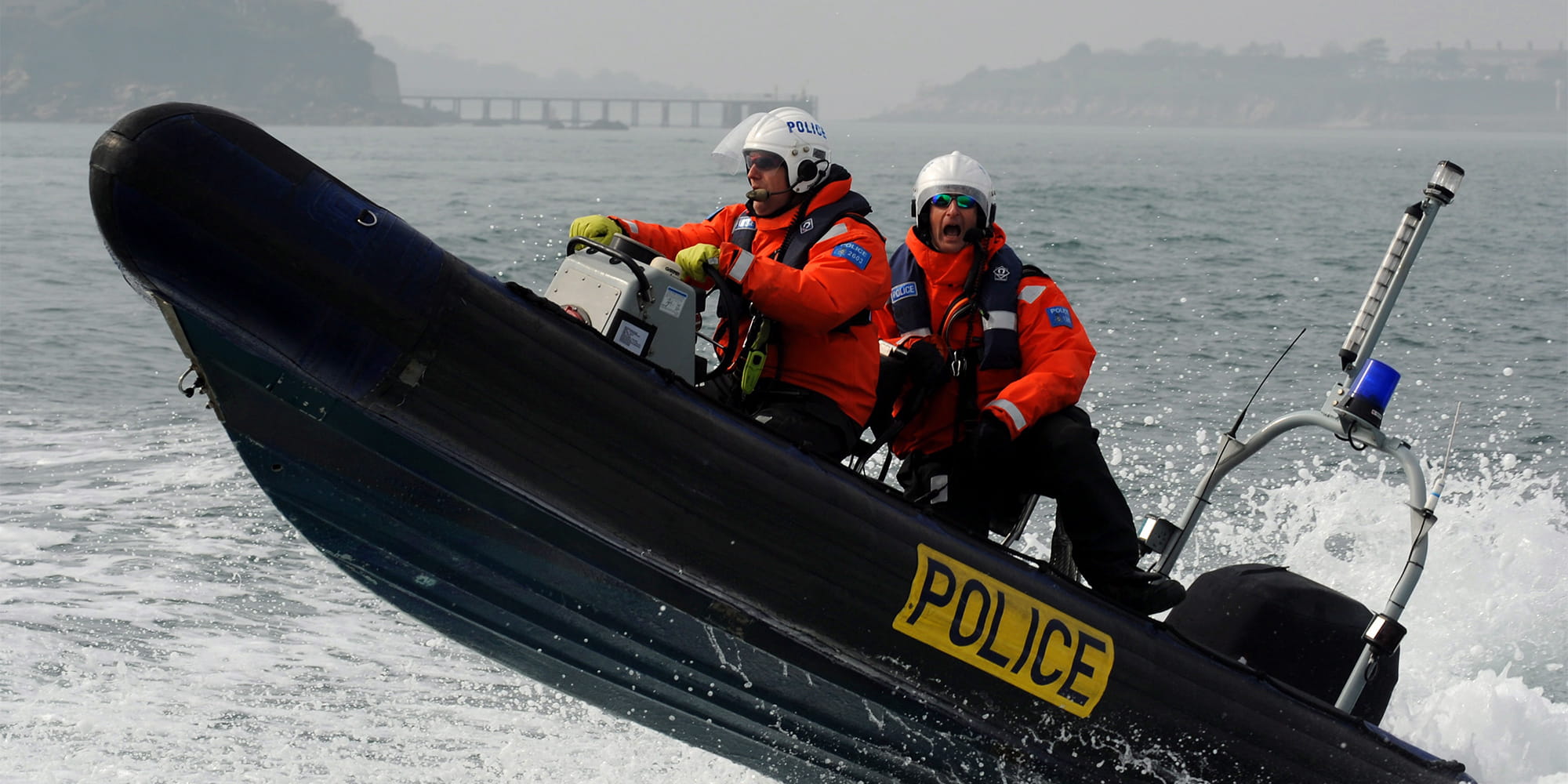2 individuals on a MoD marine police unit rib boat in the sea with its front side raised in the air after the boat hit a wave