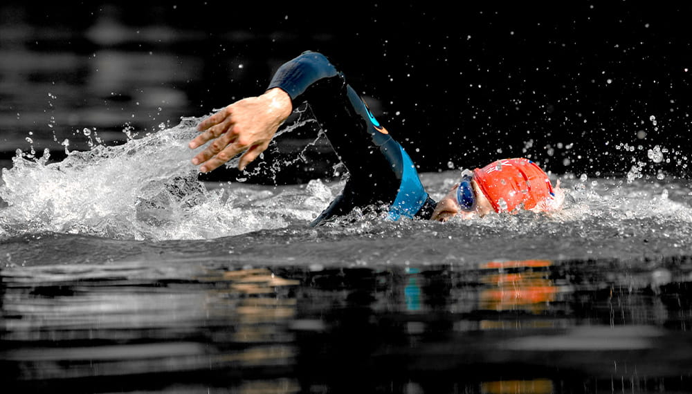 An individual in a red swim cap swims front crawl in dark water