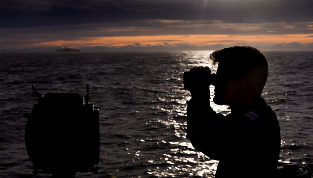 Navy sailor on a boat at sea glancing through binocular at dusk