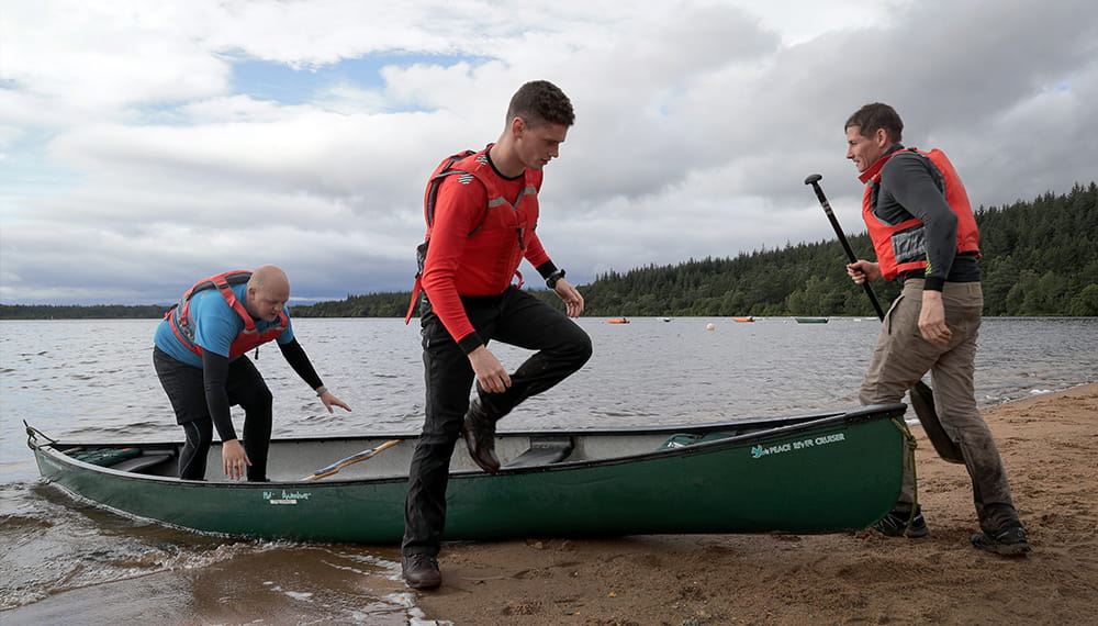 3 individuals disembark off of a large canoe on shore.