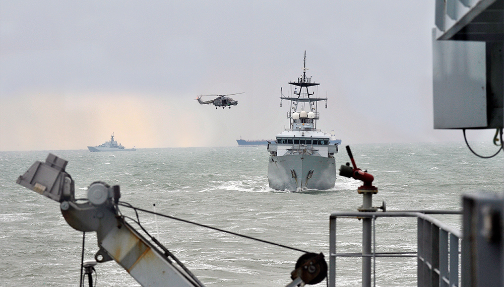 A hunt class boat approaching the camera with a helicopter circling it