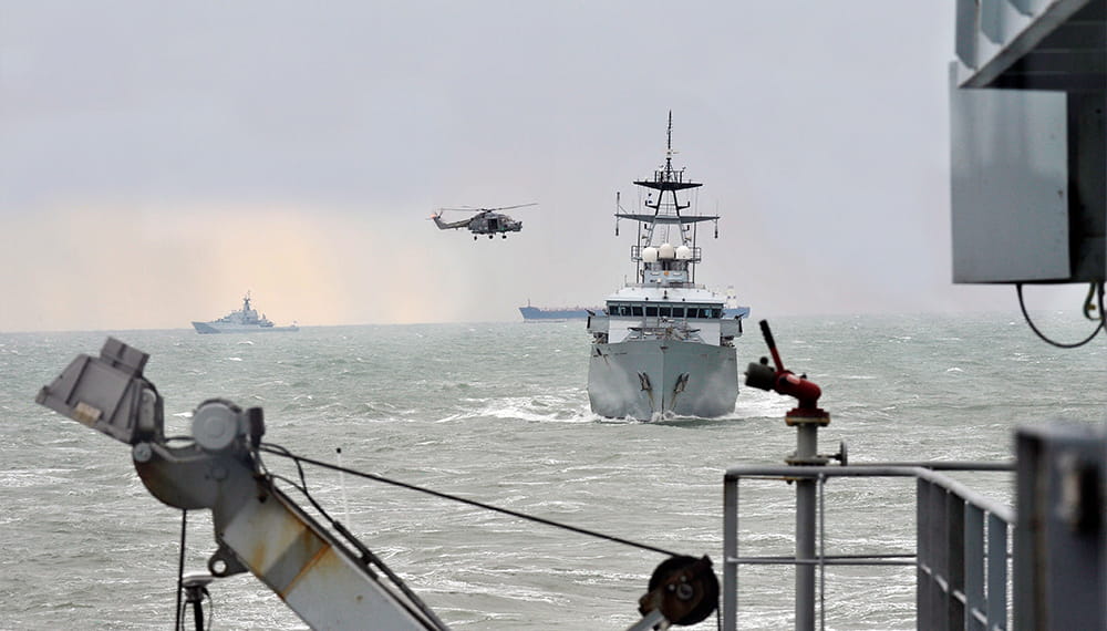 A hunt class boat approaching the camera with a helicopter circling it