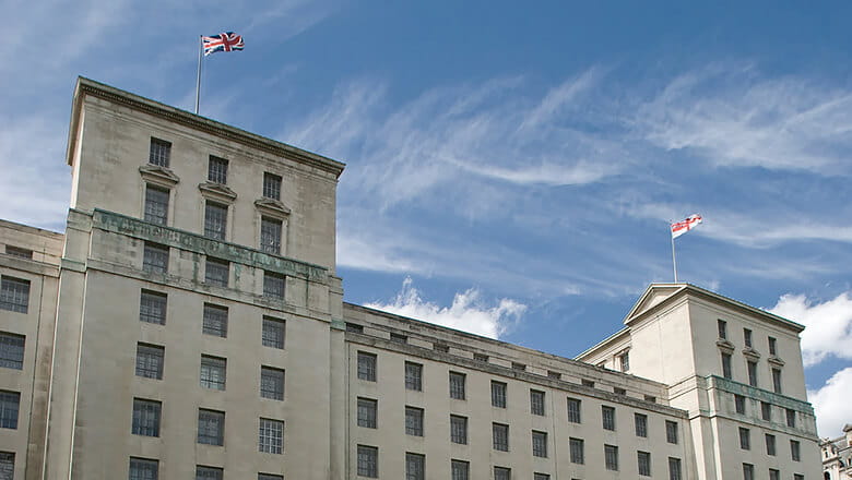 A low-angle colour photo shows the main buiding exterior of the Ministry of Defence in Westminster. A Union Jack flag and a Royal Navy White Ensign flag fly high atop the stone structure.