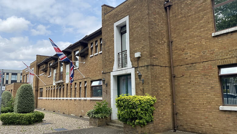 A colour photo shows the main exterior building of the Royal Marine Reserve unit in Wandsworth, London. The brick building is accented by a Union Jack flag and a Royal Navy White Ensign flag.