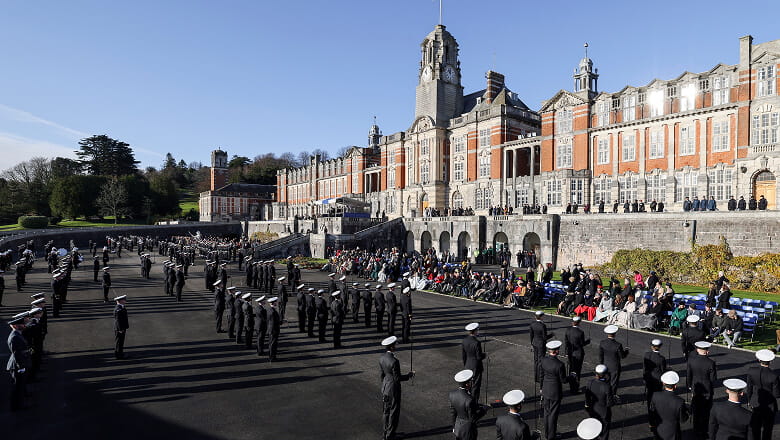 A colour photo shows the main exterior building and courtyard of the grand Brittania Royal Naval College. Service members and families gather together in the large courtyard for a passing out ceremony.