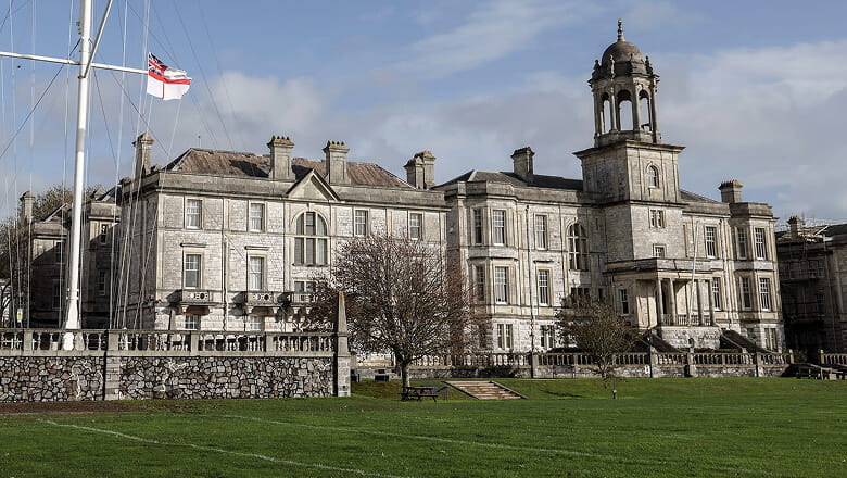 A colour photo shows the main exterior building of His Majesty’s Naval Base Devonport in Plymouth. The grand building stands before a large plain of green grass.
