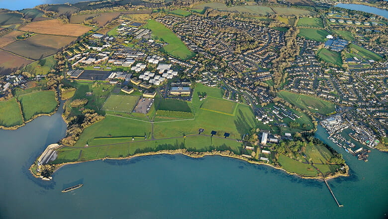 A colour photo shows a bird's eye view of the HMS Raleigh base. The vast estate features fields of grass and several large clusters of buildings.