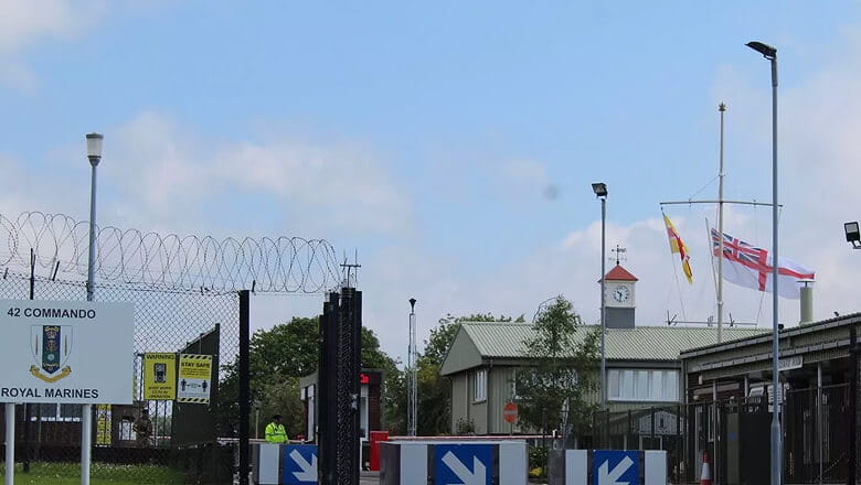 A colour photo shows a cropped view of the entrance to the Royal Marines 42 Commando base. Several traffic and safety signs mark the entrance gate and surrounding structures. A Royal Navy White Ensign flag and an RM 42 Commando fly near the entrance.