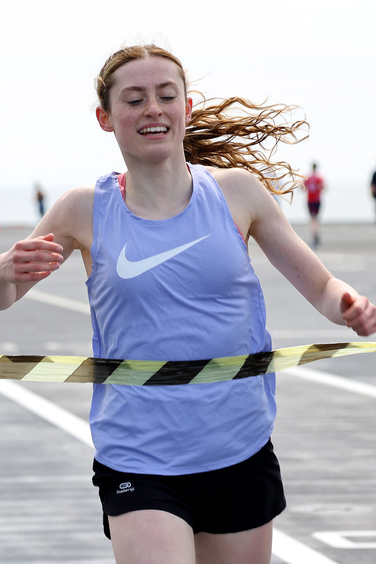 A woman is wearing a blue vest and crossing a finish line in a race