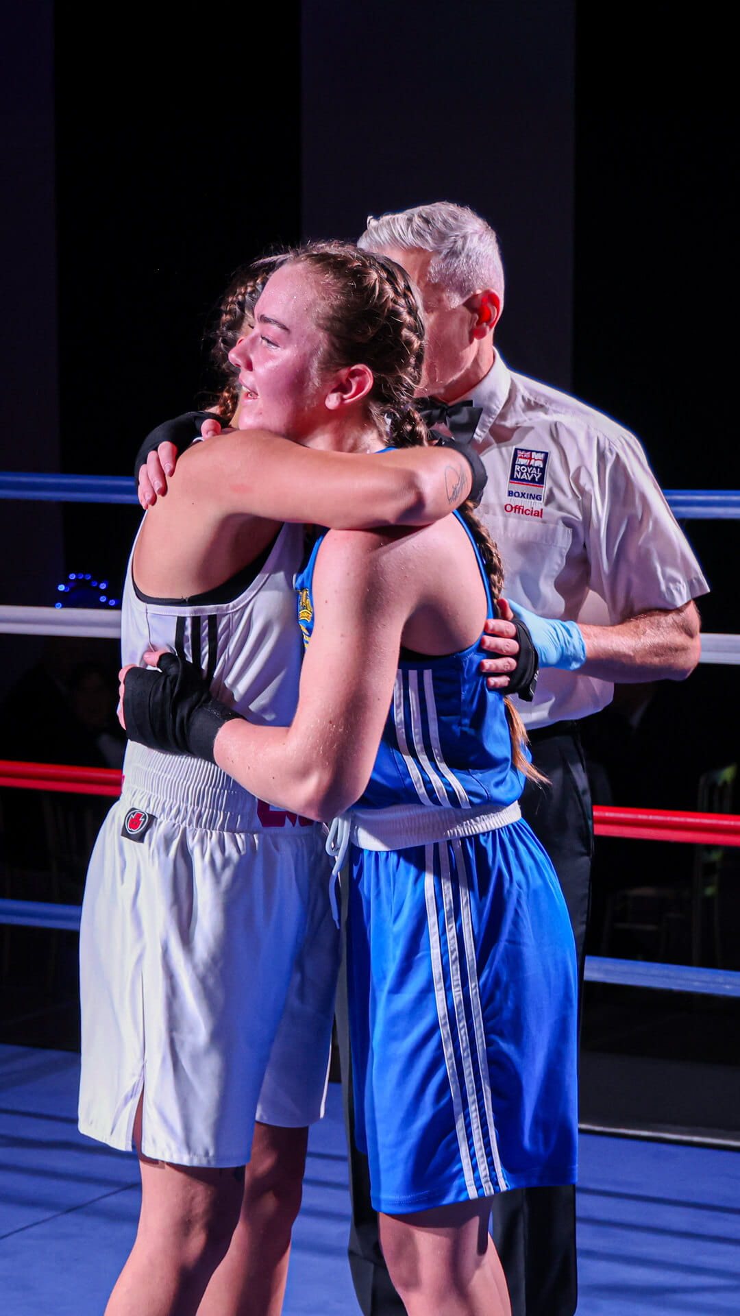 Two Royal Navy women's boxers embrace in the ring at the end of a match. A Royal Navy referee stands behind them.