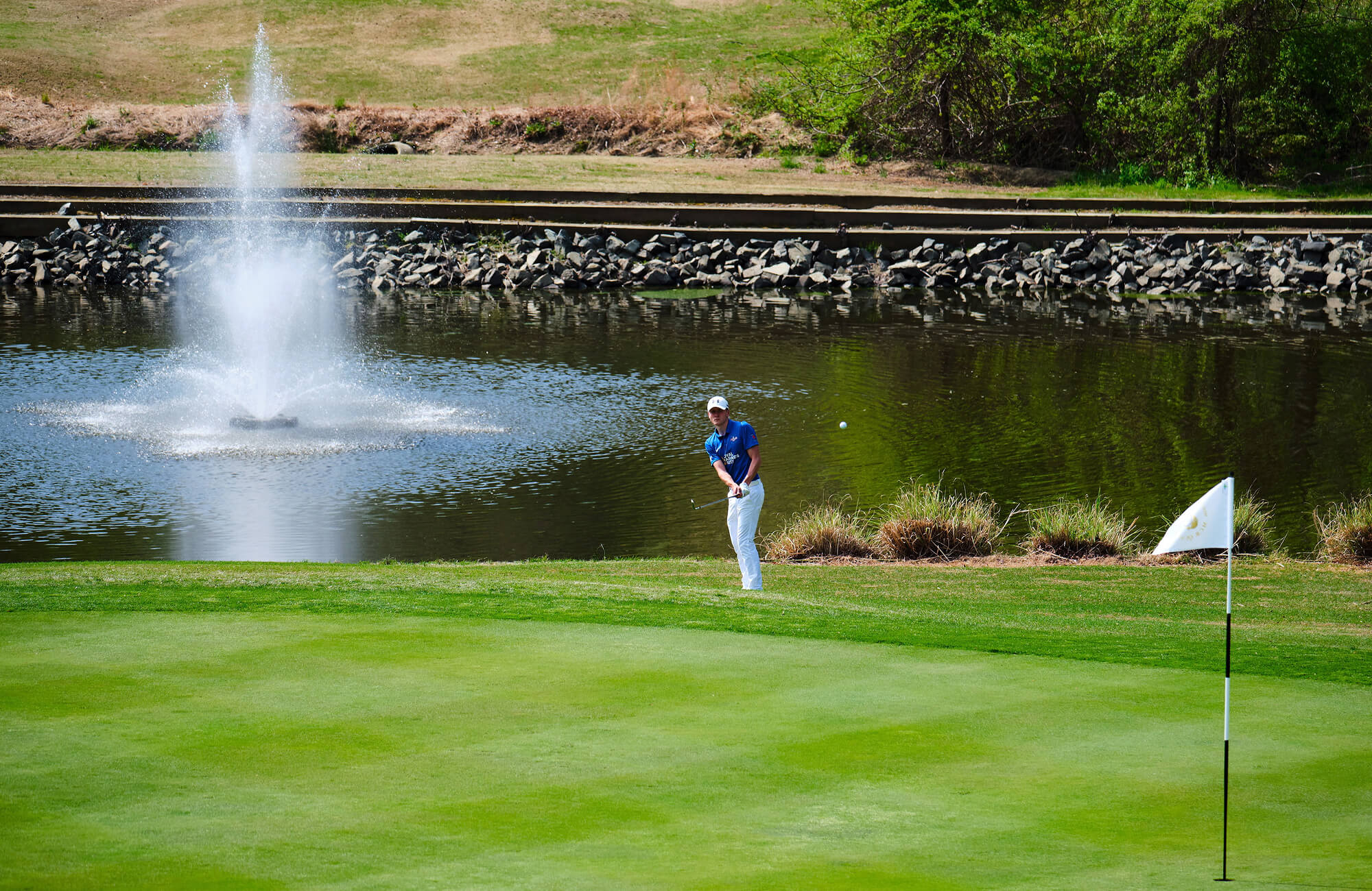 A colour image shows a wide angle of a Royal Navy golfer on course. He is swinging a club, as a ball heads towards a hole in the foreground. In the background is a large water fountain and pond.