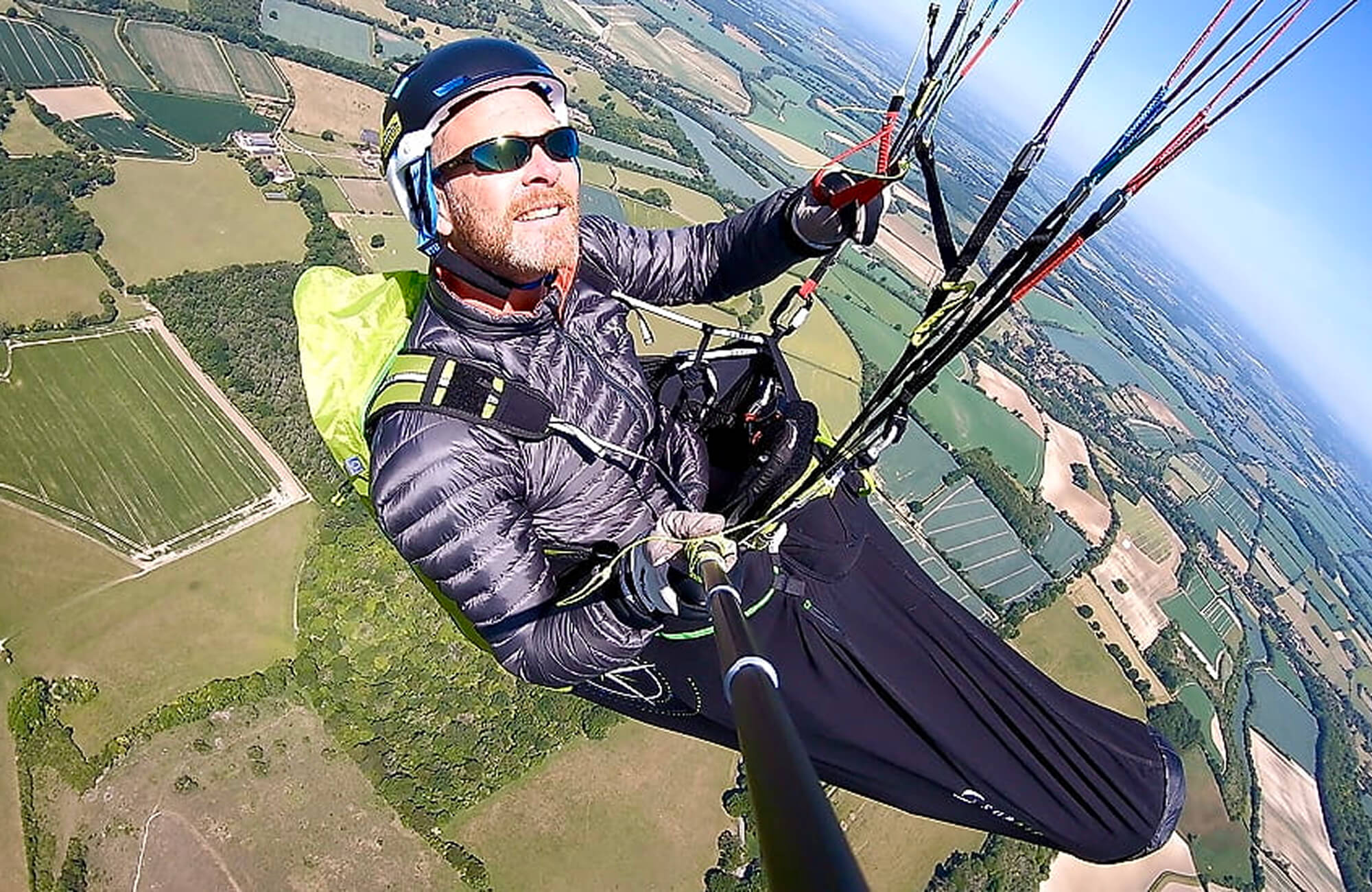 A colour photo shows a close-up, birdseye view of a Royal Navy paraglider flying above a green countryside. He looks toward the camera as the sun shines brightly in the distance. In the distance, the horizon of the earth is clearly visible.