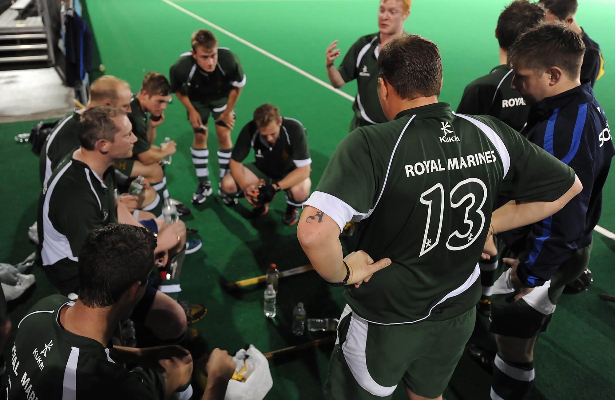 A colour photo shows a Royal Navy men's hockey team huddled on the sidelines of a field. They wear official Royal Navy hockey kit and appear to be discussing gameplay.