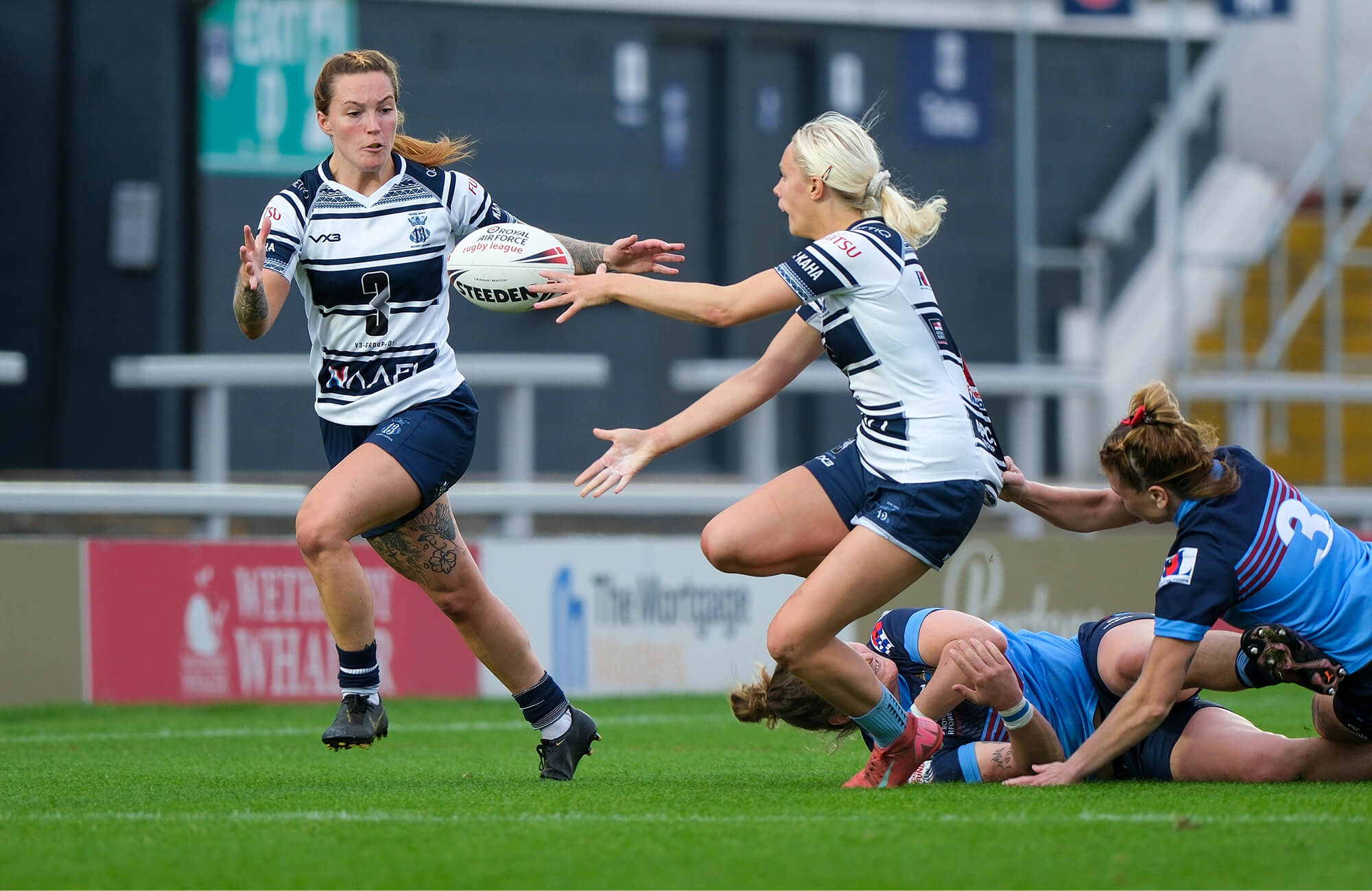 A colour photo shows two Royal Navy women's rugby league players passing the ball to one another. In the background, opponents from the competing team tumble to the ground.