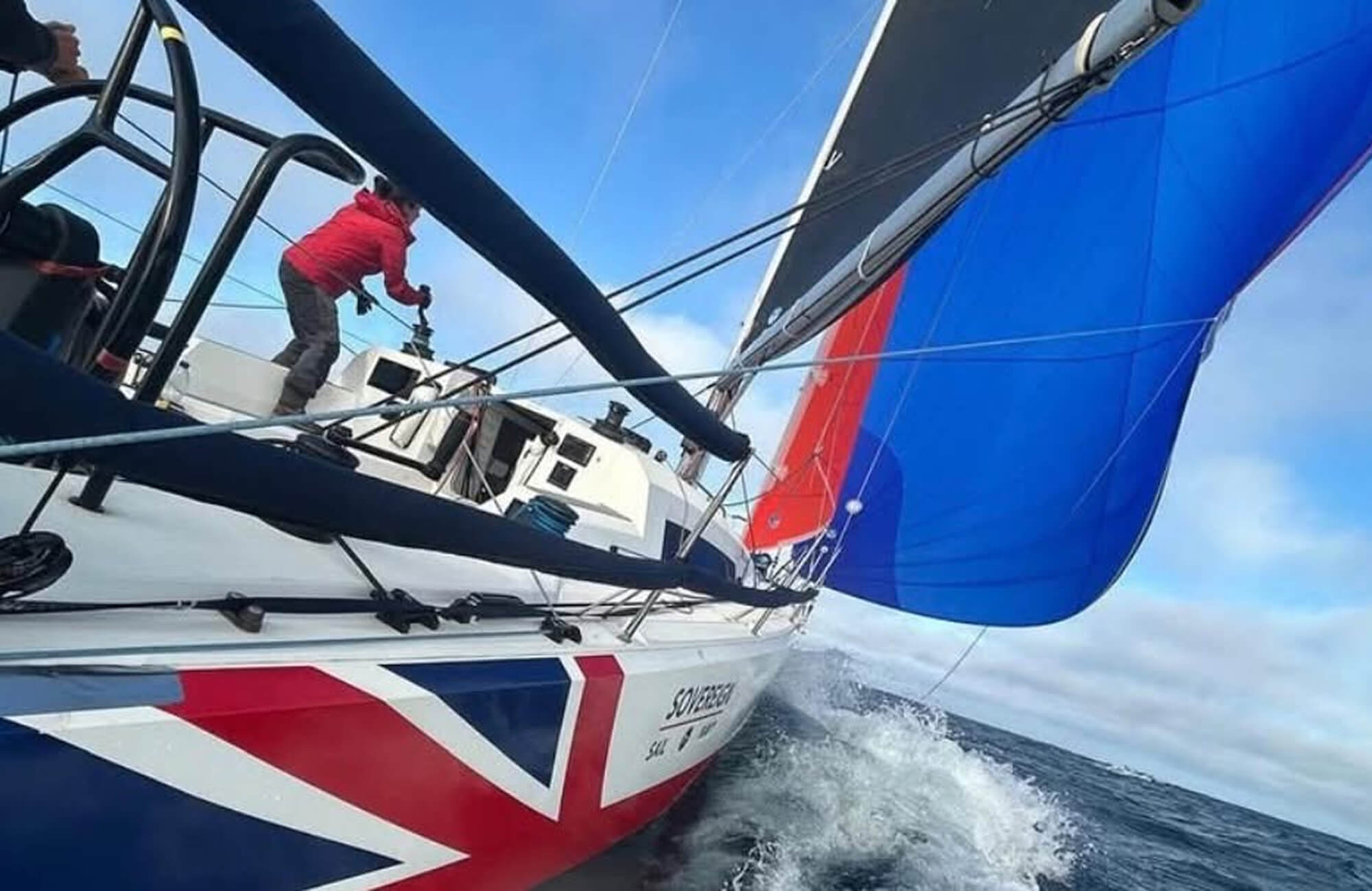A dynamic colour photo shows a Royal Navy yacht racing through open waters. Its hull is painted in a Union Jack decal. One sailor mans the controls as their crew mate controls the sails. The water foams and splashes as the boat speeds through.