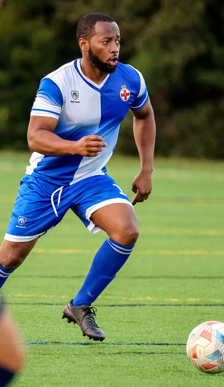 A colour photo shows a Royal Navy footballer on the pitch, mid-stride. Just ahead of his feet is a football. He wears official Royal Navy football kit.