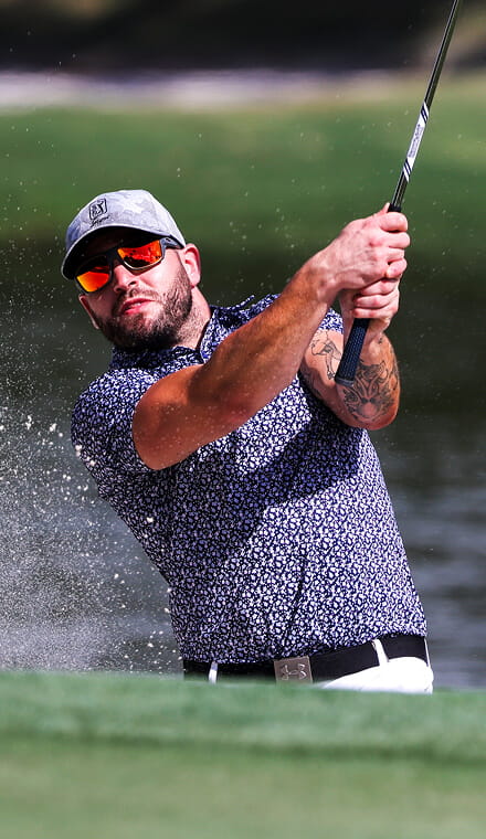 A colour photo shows a Royal Navy golfer as he swings a golf club overhead. He wears reflective sunglasses and a baseball cap. In the foreground, particles of sand are visible. In the blurred background, a lake is visible but out-of-focus.