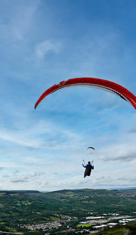 A colour photo shows a Royal Navy hang glider flying above a hilly town with a red parachute. The landscape below is lush and green, with clusters of buildings in between.