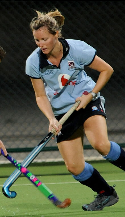 A colour photo shows a Royal Navy hockey player on the field, mid-play. She holds a field hockey stick and uses it to block an opponent's move.