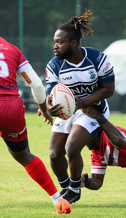 A colour photo shows a Royal Navy rugby league player on the pitch. He holds a rugby ball close to his chest, while an opponent dives to tackle him.