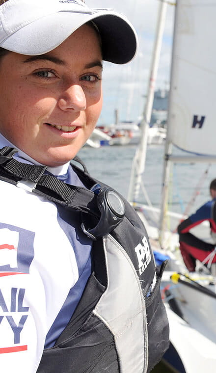 A colour photo shows a Royal Navy keelboat sailor smiling at the camera. She wears a baseball cap and safety gear. A white boat sail is visible in the background.